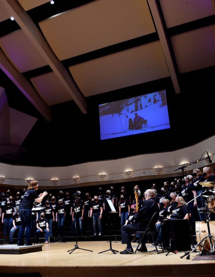 A choir performs on stage with a conductor leading, musicians seated nearby