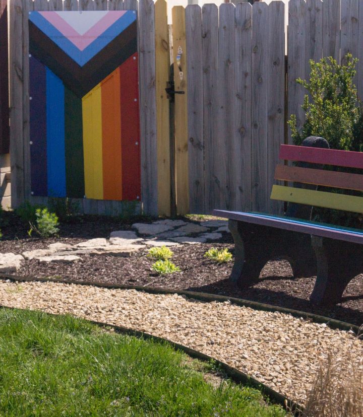 An outdoor garden space with a gravel path, a rainbow-colored bench, and a Progress Pride flag mural on a wooden fence.