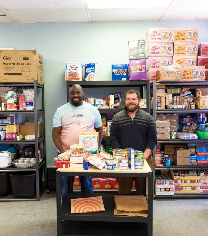 Two men stand smiling behind a table of food donations in a well-stocked food pantry.